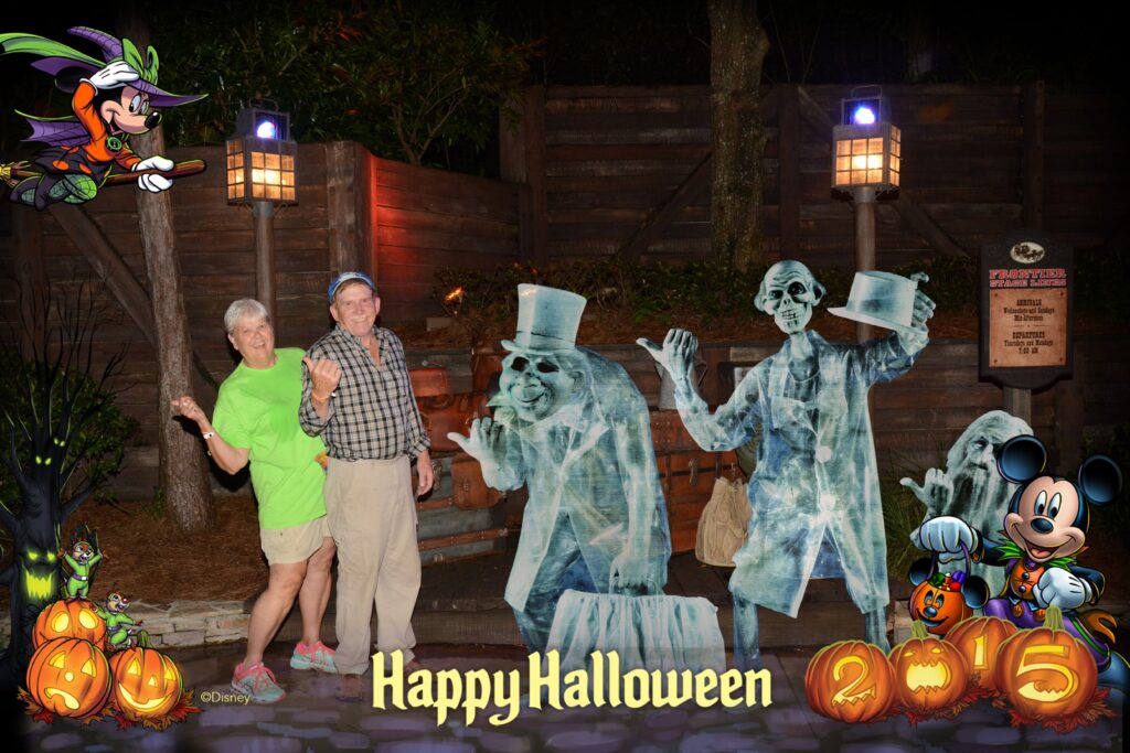 PhotoPass_Visiting_Mickeys_Not_So_Scary_Halloween_Party_7483127584 A smiling couple poses at night beside Disney’s animated Hitchhiking Ghosts from the Haunted Mansion during Mickey’s Not-So-Scary Halloween Party. The photo is framed with Halloween decorations, including pumpkins, Minnie Mouse flying on a broomstick, and Mickey Mouse dressed as a vampire with the text “Happy Halloween 2015.”