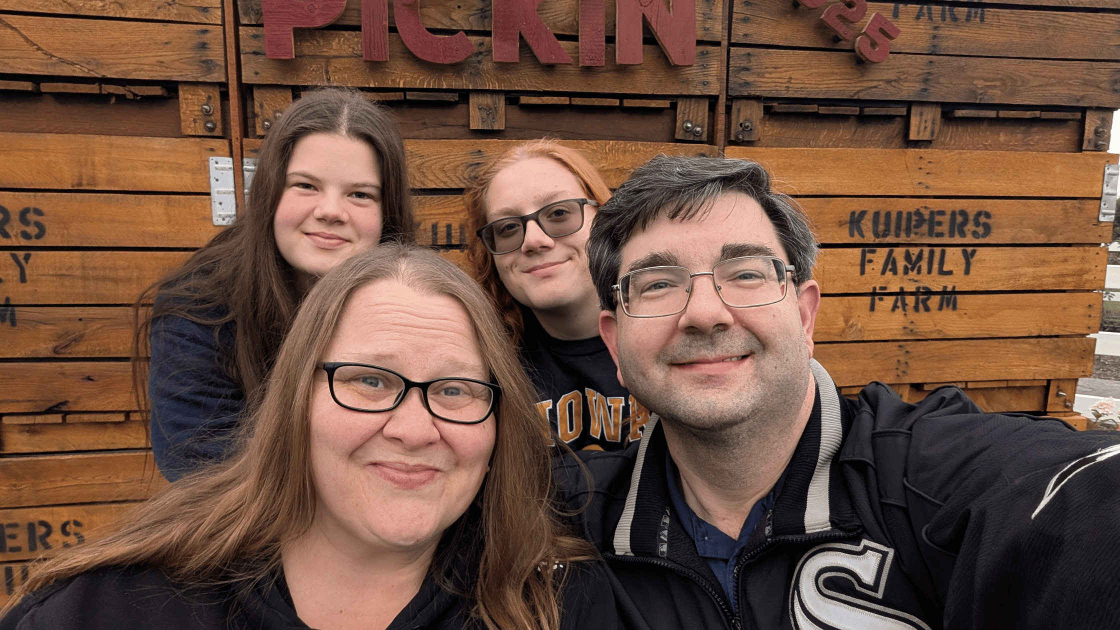 A family of four smiling together in front of wooden crates marked “Kuipers Family Farm,” enjoying a cozy day and capturing a happy moment together.