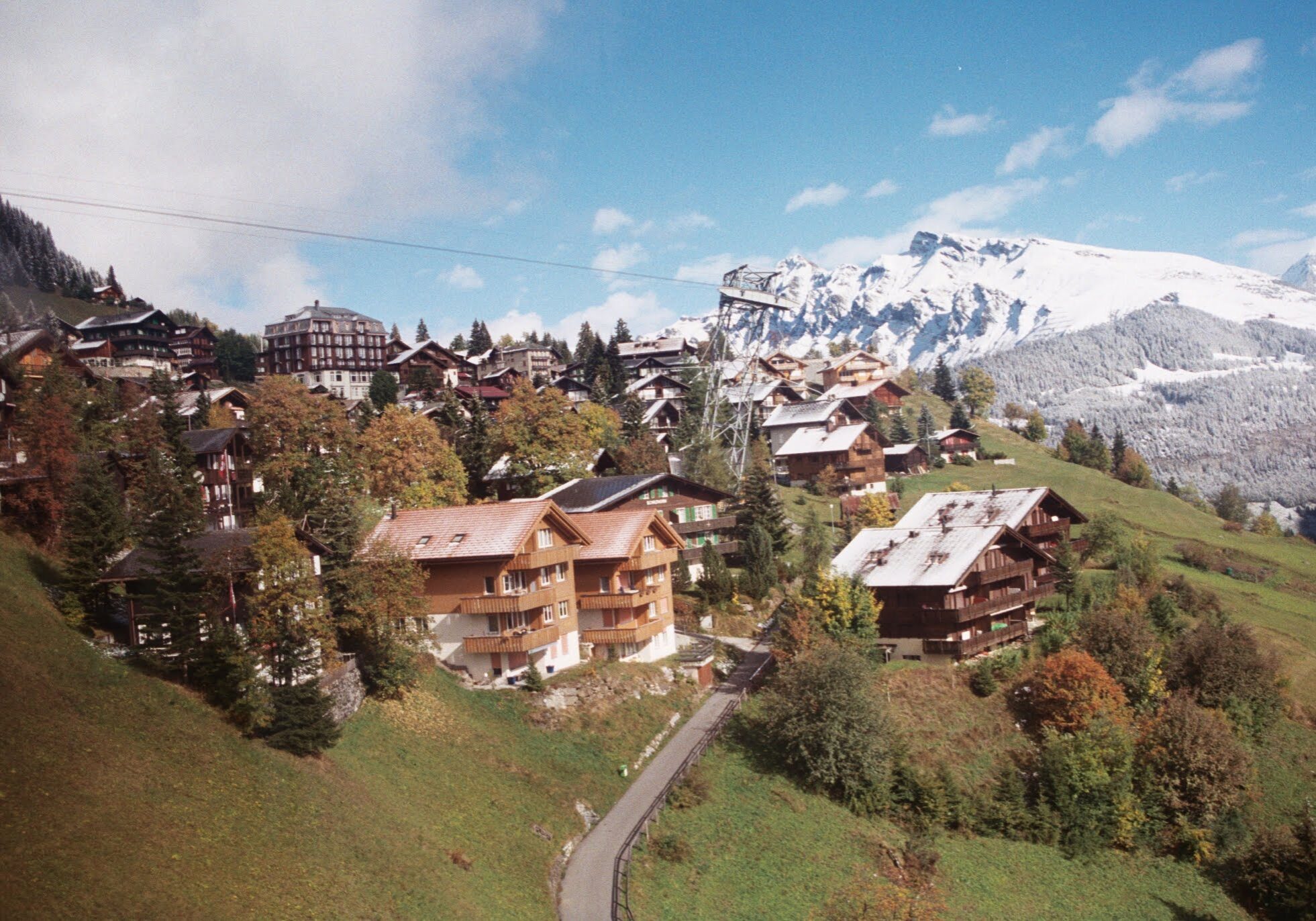 A scenic mountain village with wooden chalets scattered along a green hillside, dusted with early snow. In the background, tall snow-covered peaks rise under a blue sky with light clouds, creating a stunning contrast of autumn and winter in the Swiss Alps.