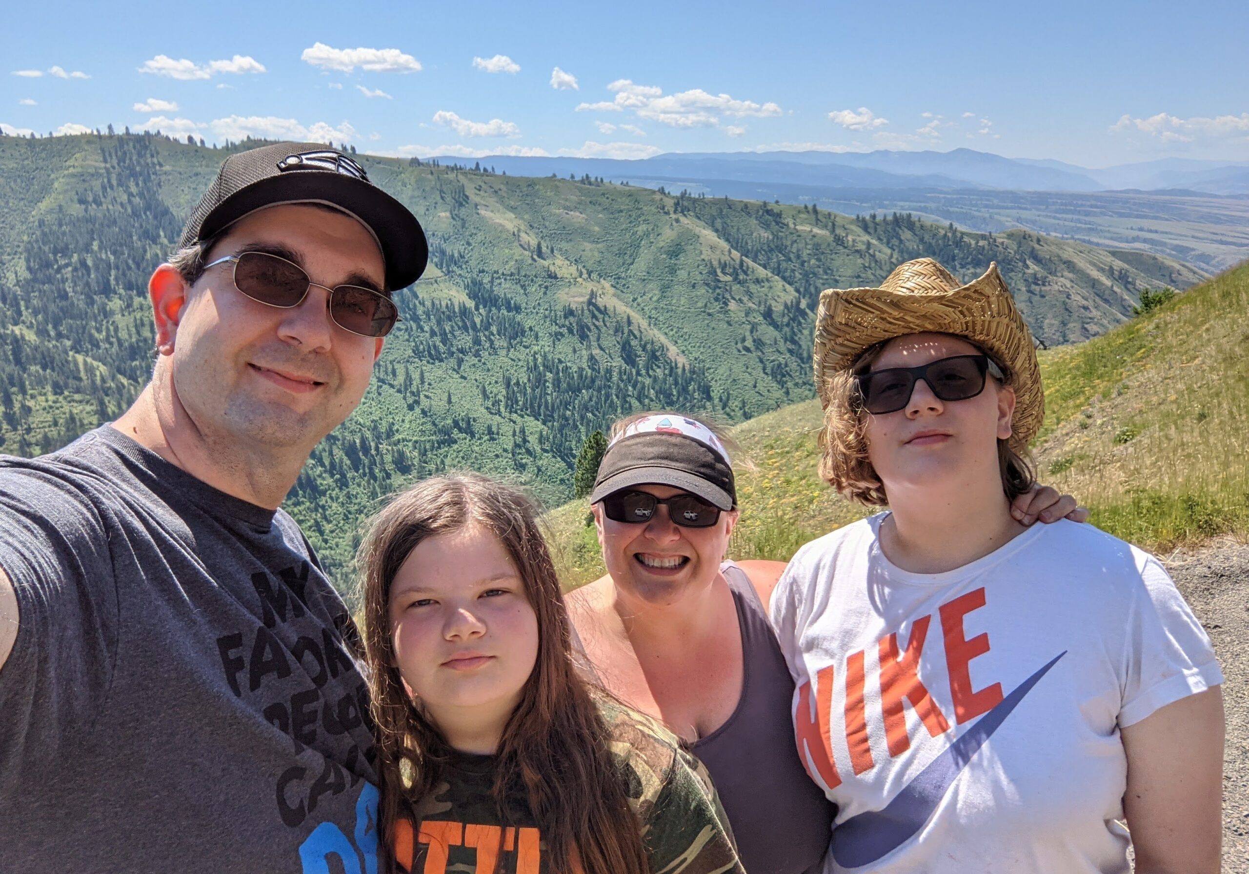 A smiling family of four takes a selfie on a sunny day with a stunning mountain valley in the background. The parents and two children wear casual summer clothes, sunglasses, and hats, standing close together on a hillside with green ridges and distant blue mountains behind them.