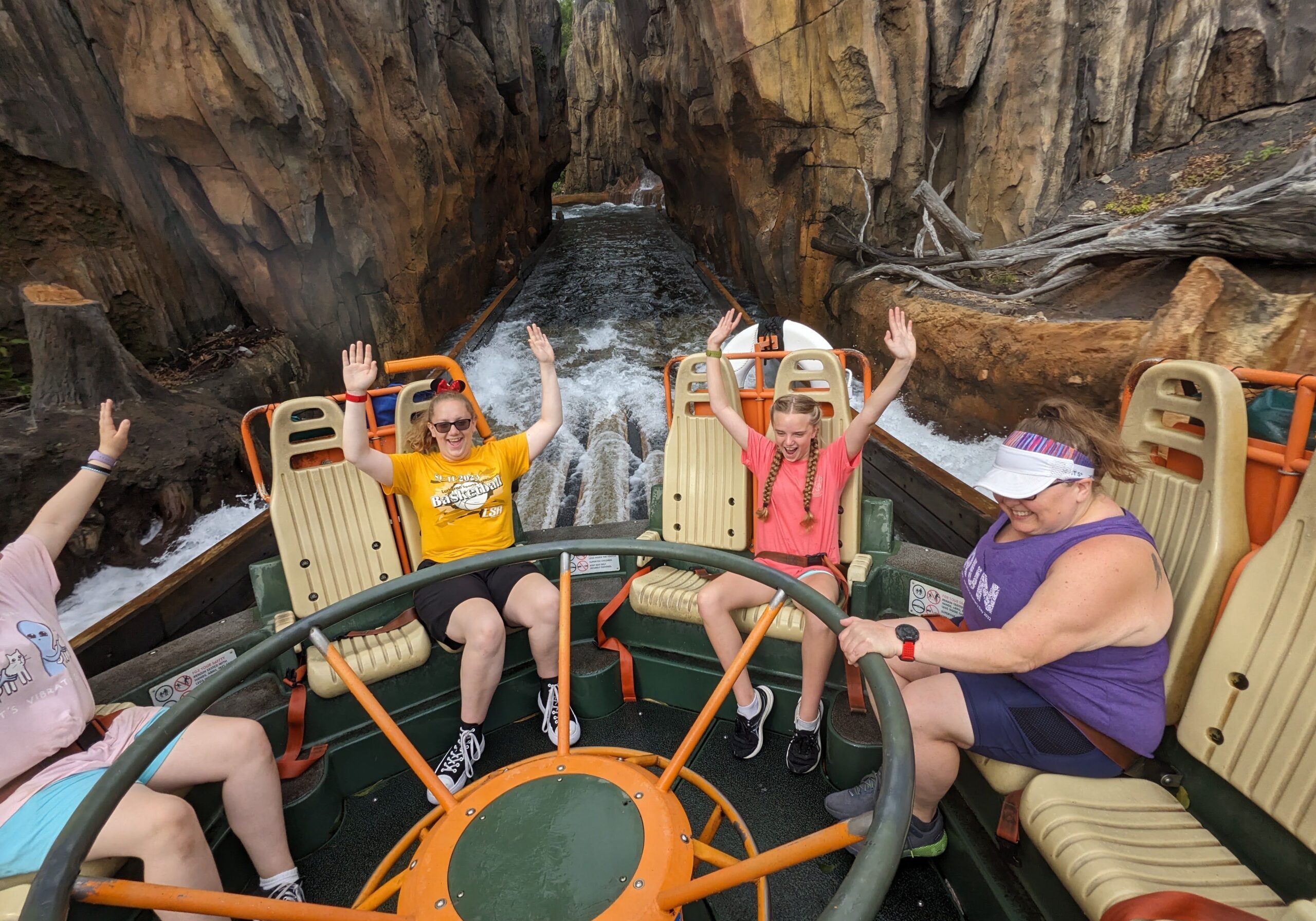 A group of four people ride a circular raft on a water rapids attraction, smiling and raising their hands as they approach a drop between rocky canyon walls. Water splashes around the raft, and everyone appears excited and joyful during the ride.